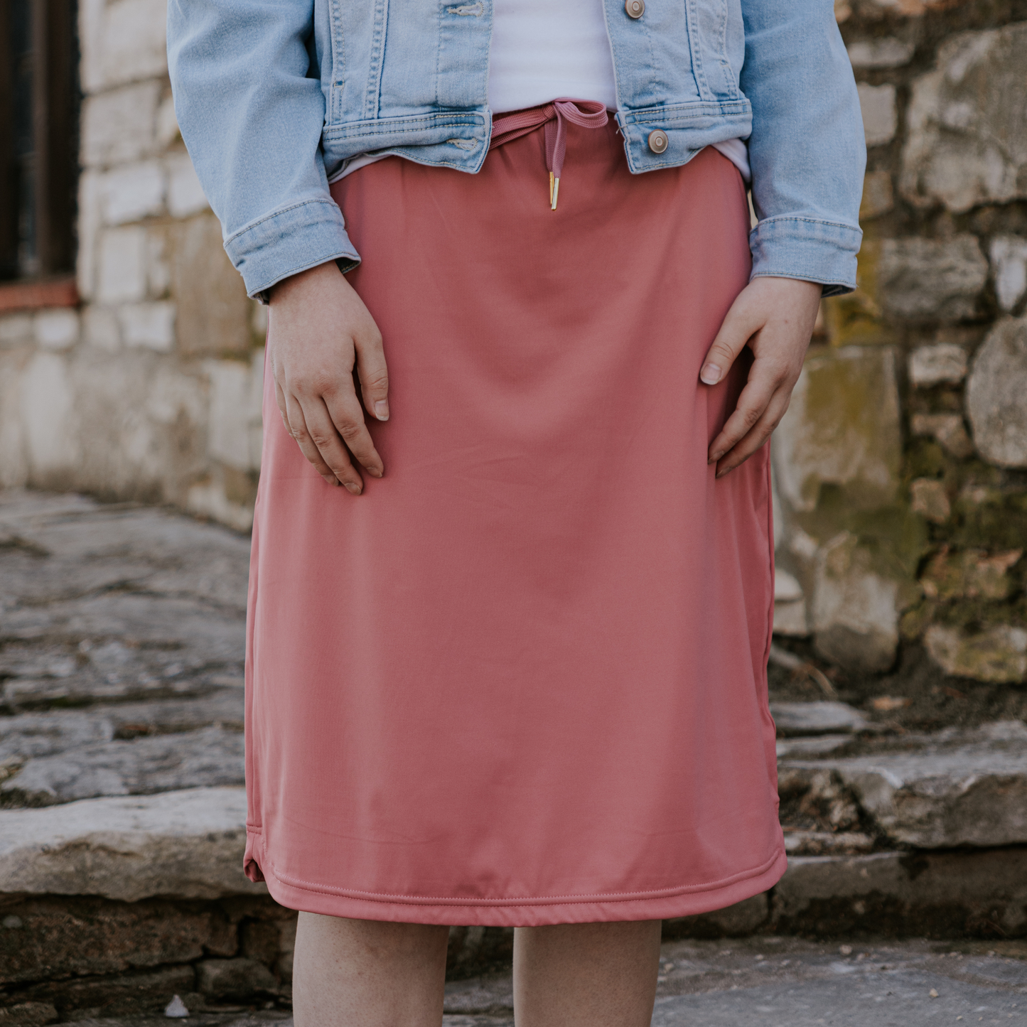 Person wearing a pink skirt and denim jacket standing against a stone wall.