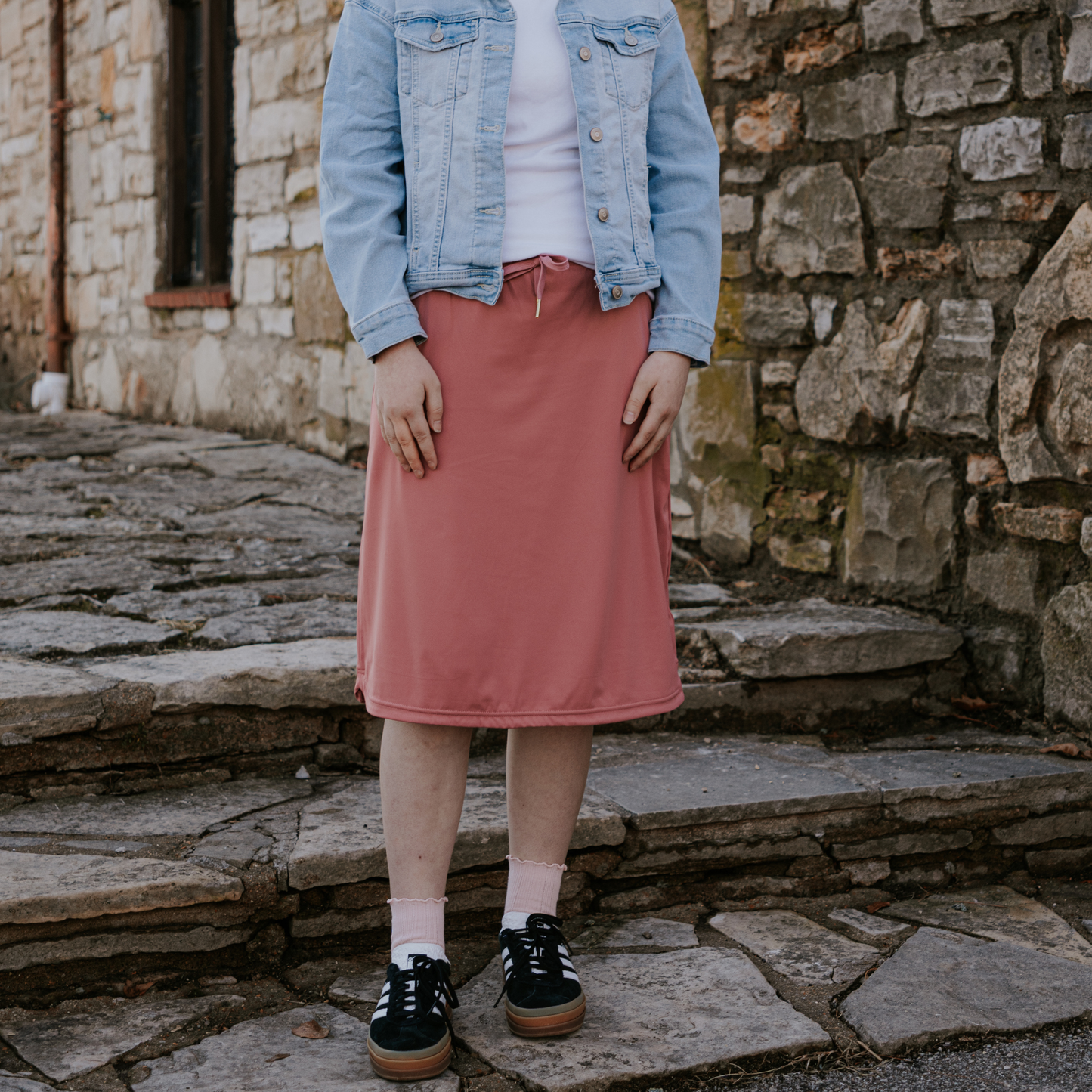 Person wearing a denim jacket and pink skirt standing on stone steps with a stone wall background