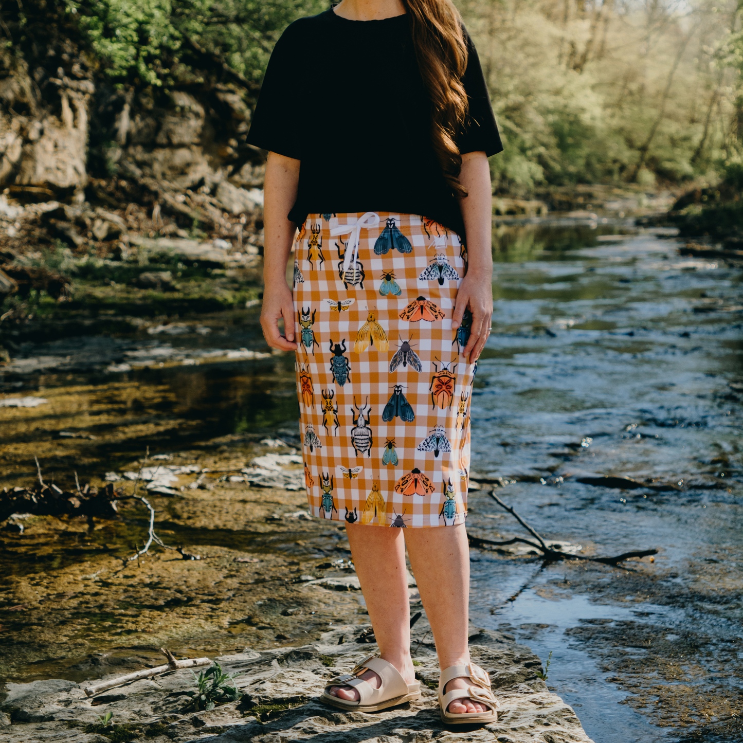 Person wearing a checkered skirt with nature-themed designs by a stream