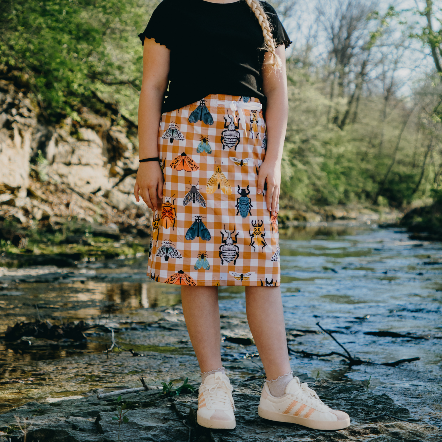 Kid wearing a checkered skirt with animal patterns standing by a stream.