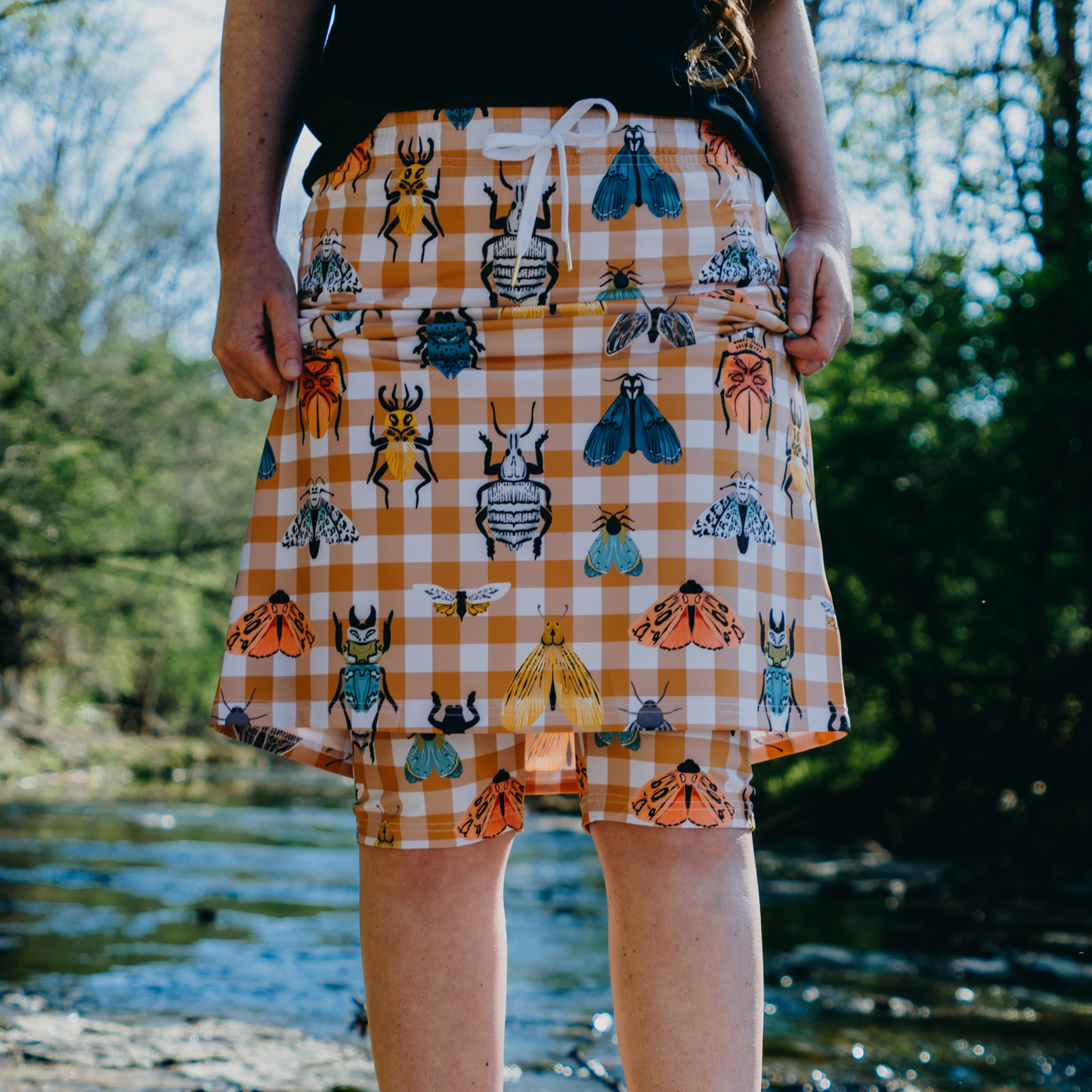 
                  
                    Person wearing a checkered skirt wit shorts with insect patterns outdoors by a body of water.
                  
                