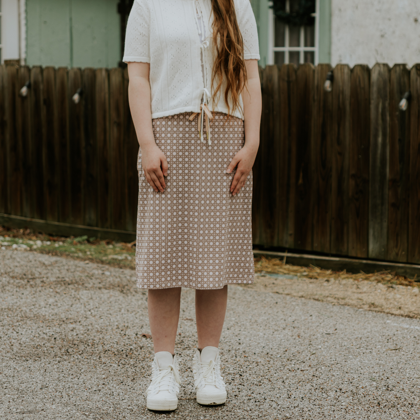 Person wearing a white shirt, patterned skirt, and white sneakers standing on a gravel surface with a wooden fence in the background.