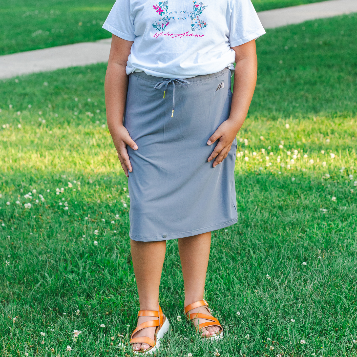 Kid wearing a light gray skirt and white t-shirt standing on grass