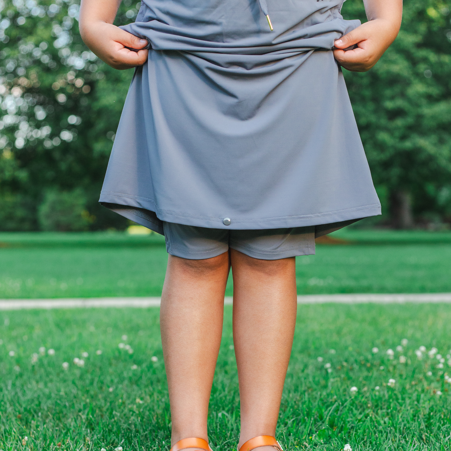 
                  
                    Kid wearing a gray skirt with shorts standing on grass with a blurred green background
                  
                