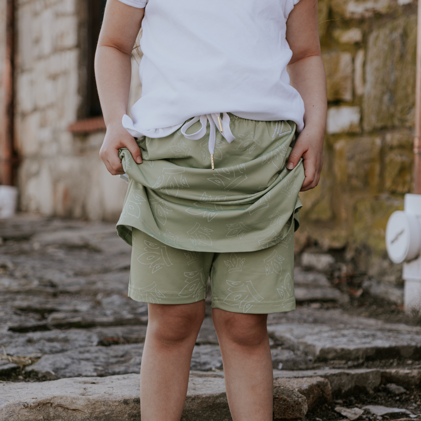 
                  
                    Person wearing green skirt and shorts and a white top in an outdoor setting with stone wall background
                  
                