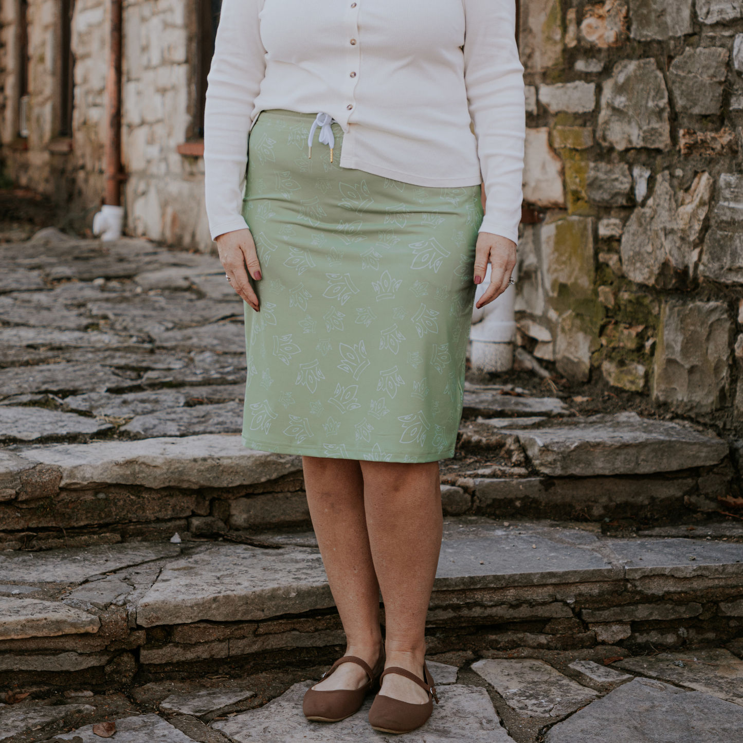 
                  
                    Person wearing a green skirt and white top standing on stone steps with a stone wall background
                  
                