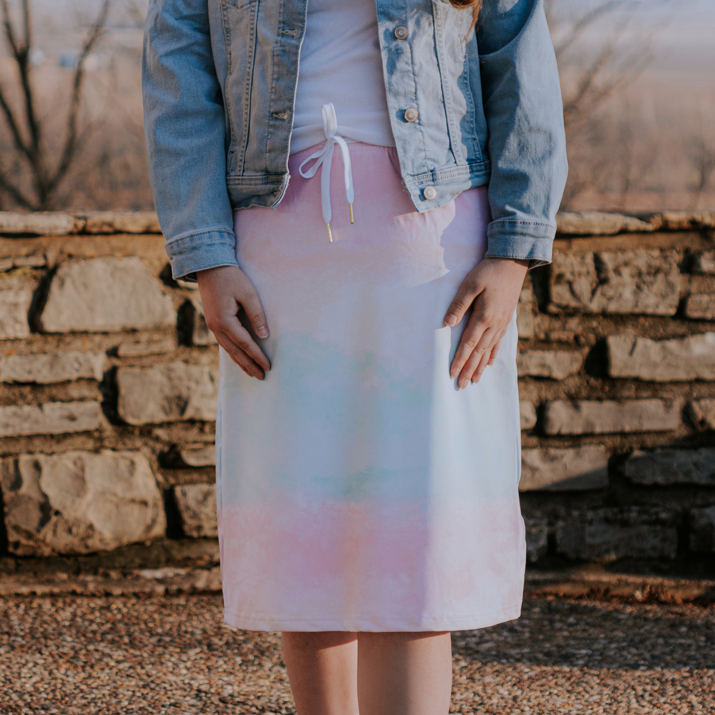 Person wearing a pastel tie-dye skirt with a denim jacket in front of a stone wall.