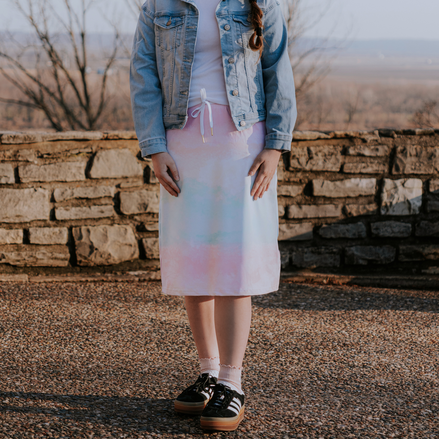 Person wearing a denim jacket and pastel skirt standing in front of a stone wall with a blurred natural background