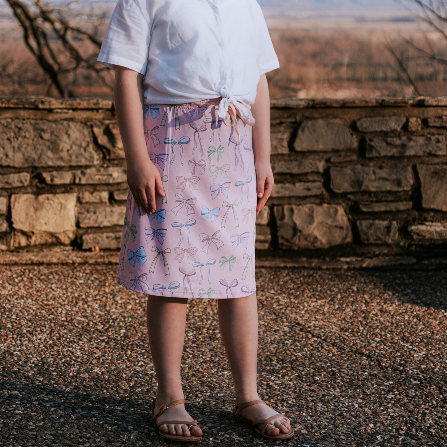 Person wearing a white top and patterned skirt standing in front of a stone wall.