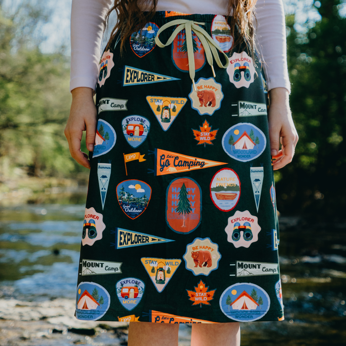 Person wearing a skirt with camping-themed patches outdoors by a lake.