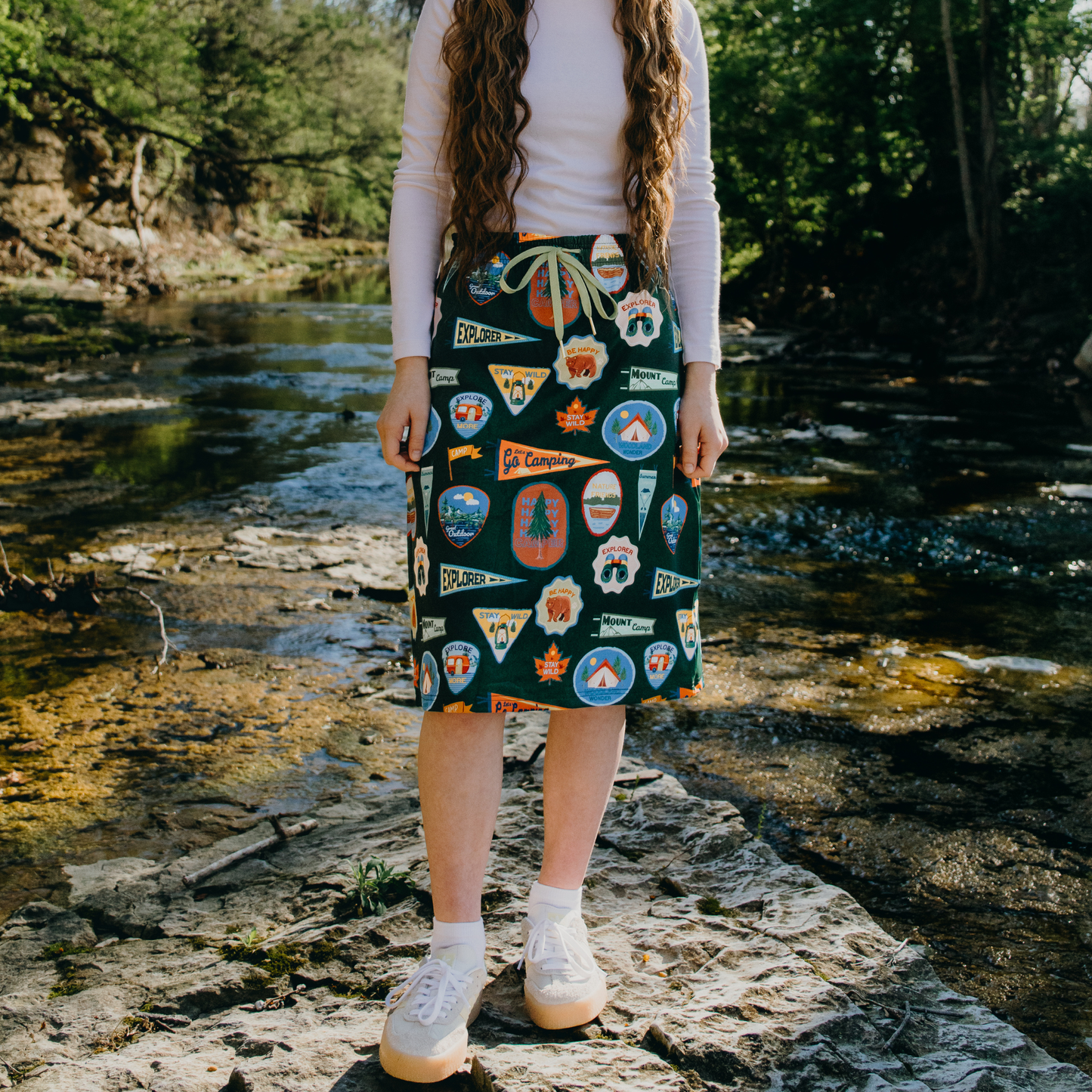 Person wearing a patterned skirt standing by a stream with trees in the background