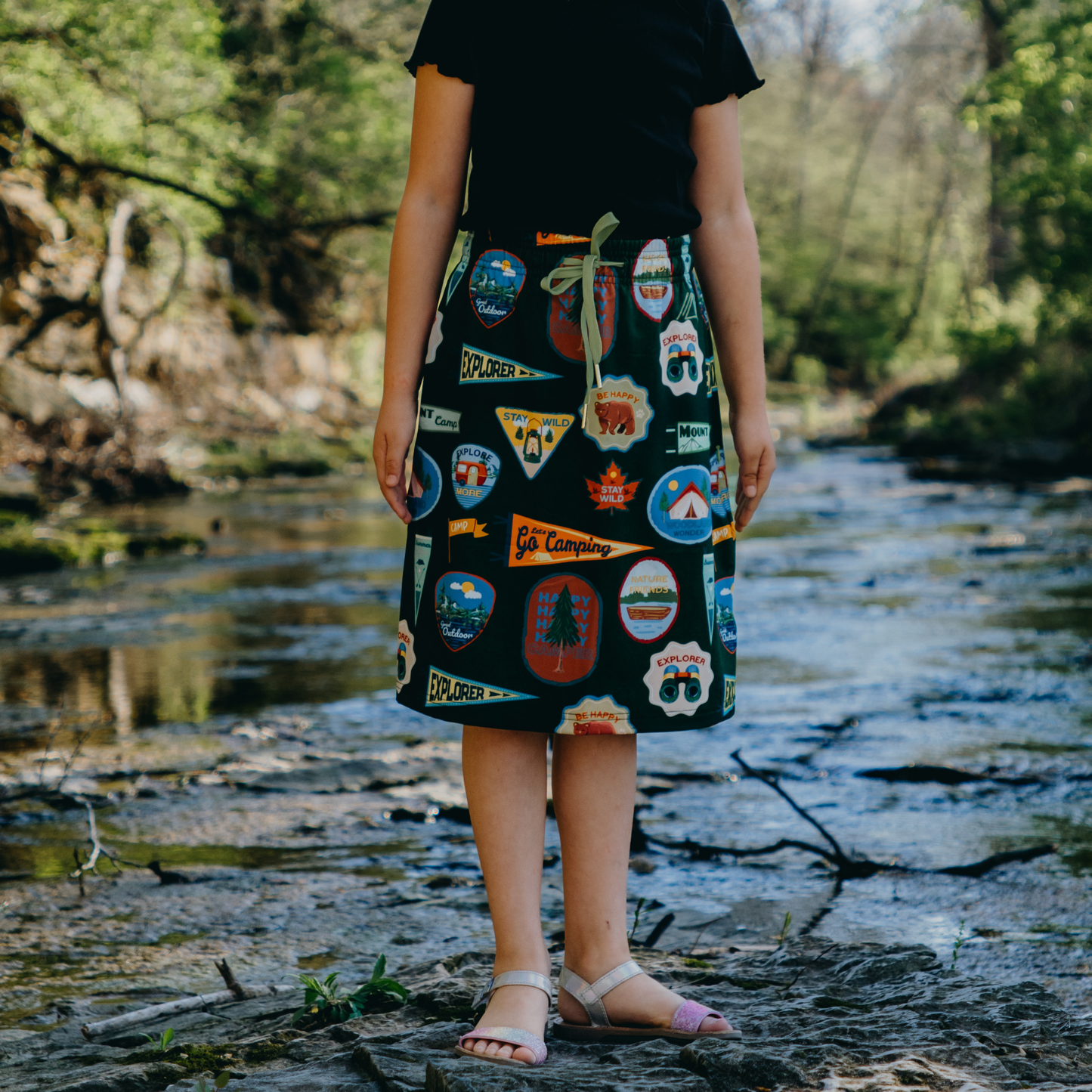 Kid wearing a skirt with camping-themed patches standing by a stream.