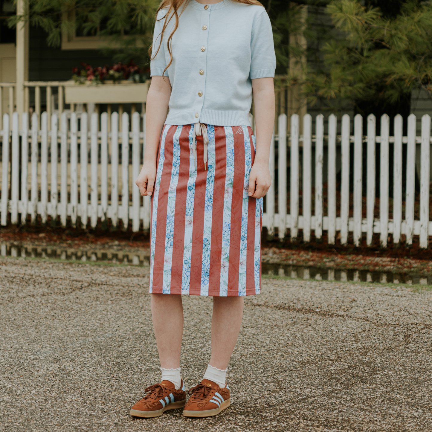 Person wearing a light blue top and red and white striped skirt standing in front of a white picket fence.