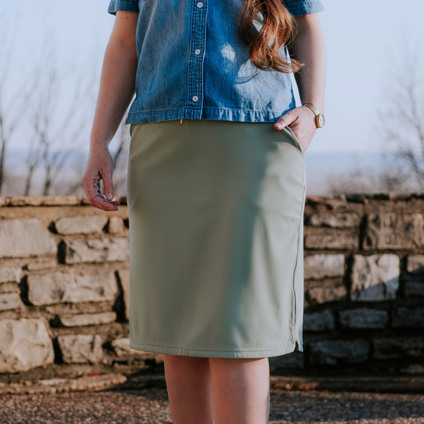 
                  
                    Person wearing a blue denim shirt and green skirt standing against a stone wall.
                  
                