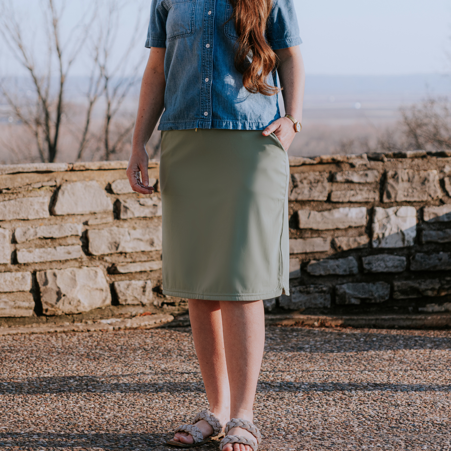 
                  
                    Person wearing a denim shirt and green skirt standing against a stone wall.
                  
                