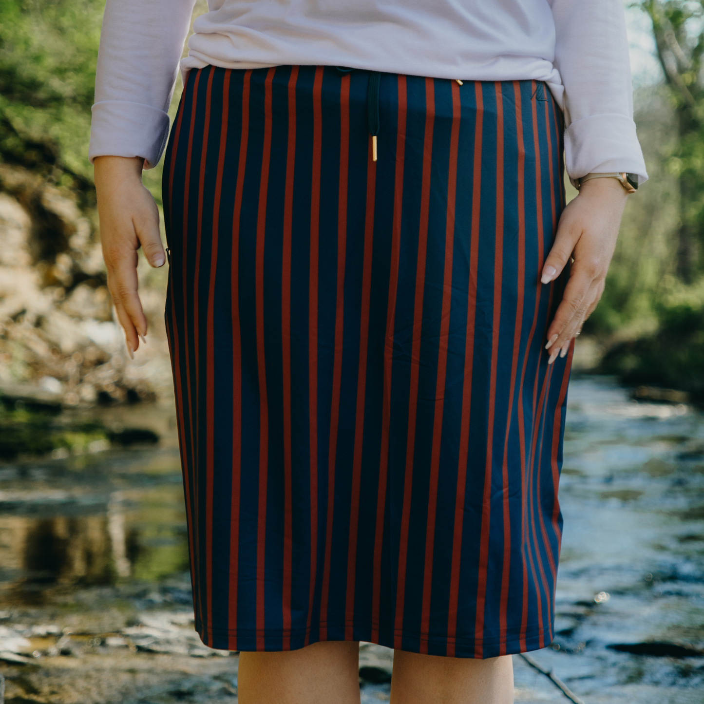 Person wearing a navy blue and orange striped skirt standing by a stream.
