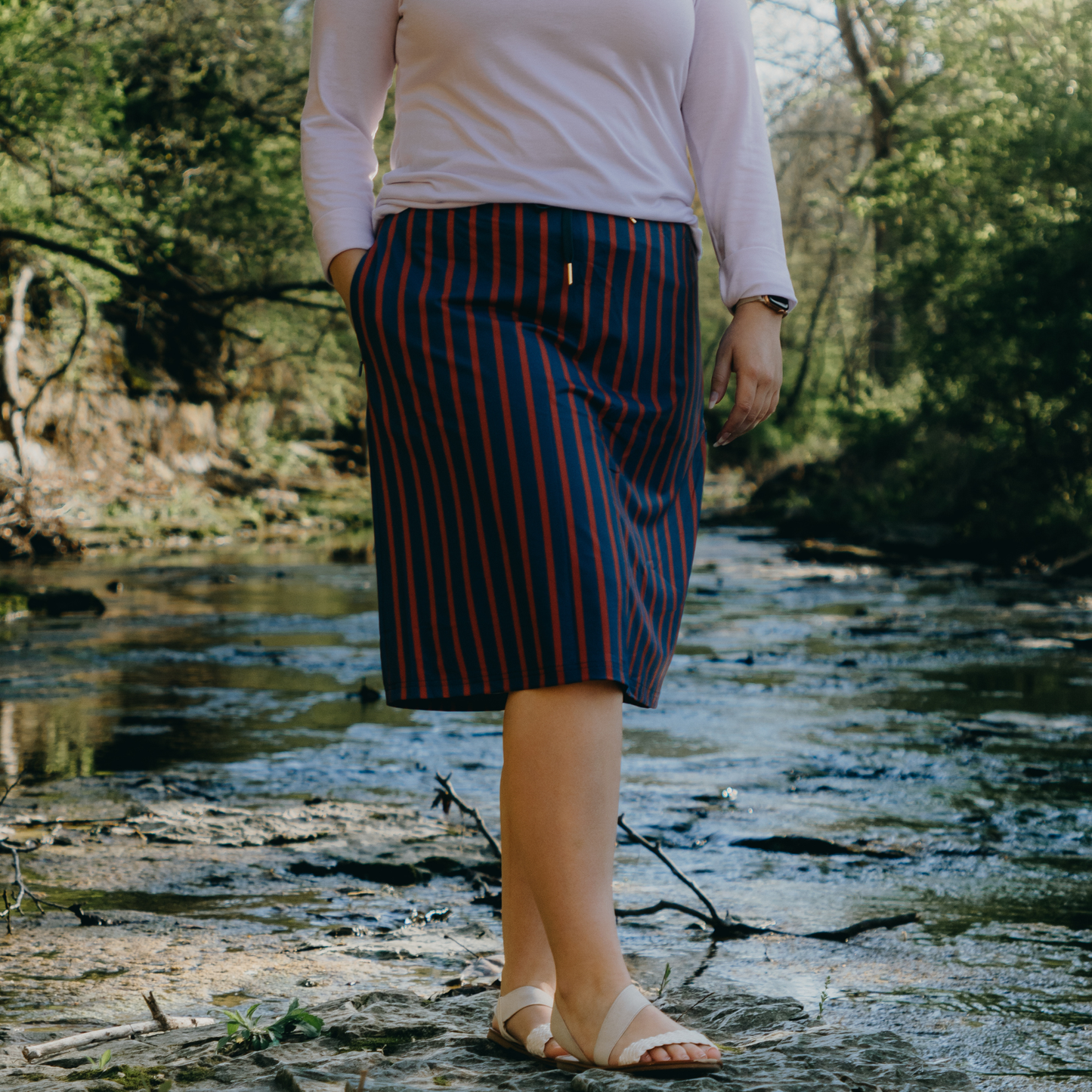 Person wearing a striped skirt standing near a stream with trees in the background