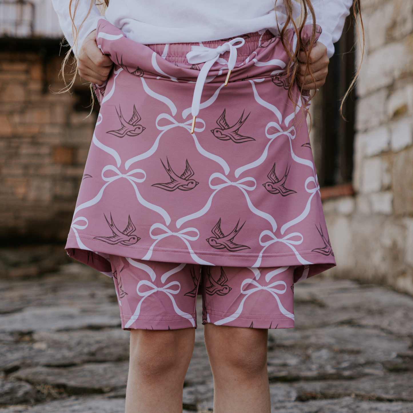 
                  
                    Child wearing pink skirt and shorts with white patterns against a stone wall background
                  
                