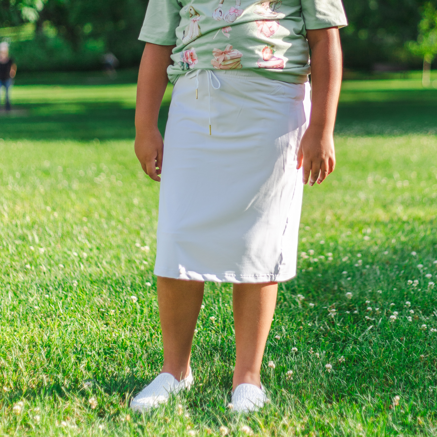 Kid wearing a green shirt with floral patterns and a white skirt standing on grass.