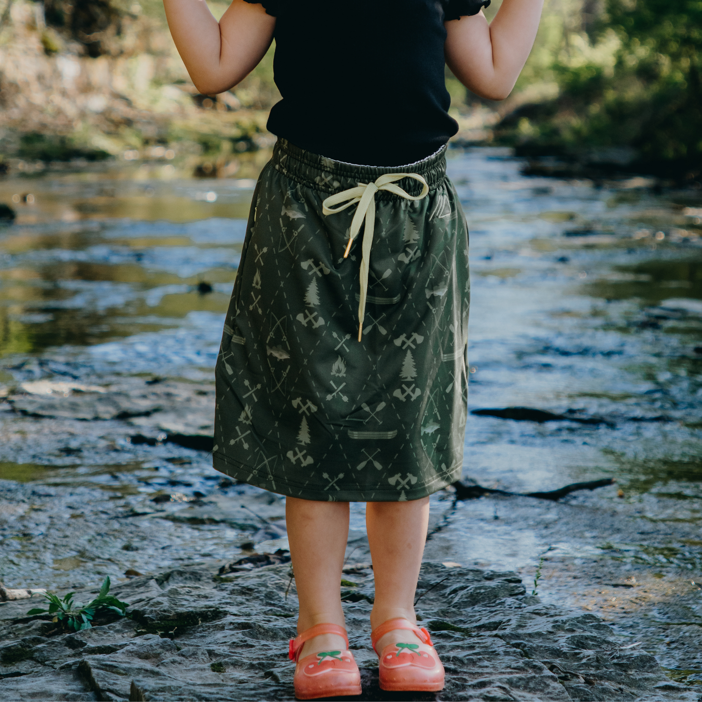 kid wearing a green patterned skirt standing near a body of water.